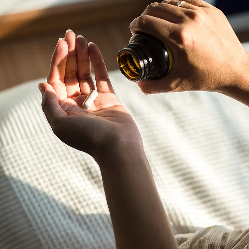 A hand holds a single white capsule while the other hand pours more from a brown medicine bottle.