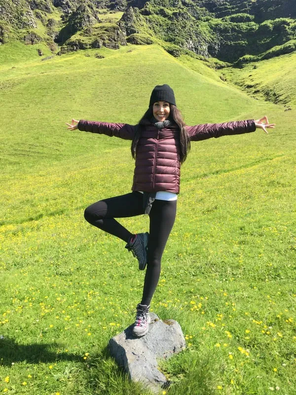 A woman in a black beanie and a maroon puffer jacket stands on a rock in a yoga tree pose with arms outstretched, set against a backdrop of a green, grassy hillside with scattered yellow flowers.