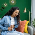 A woman clutches her stomach while sitting on a couch, showing signs of digestive discomfort