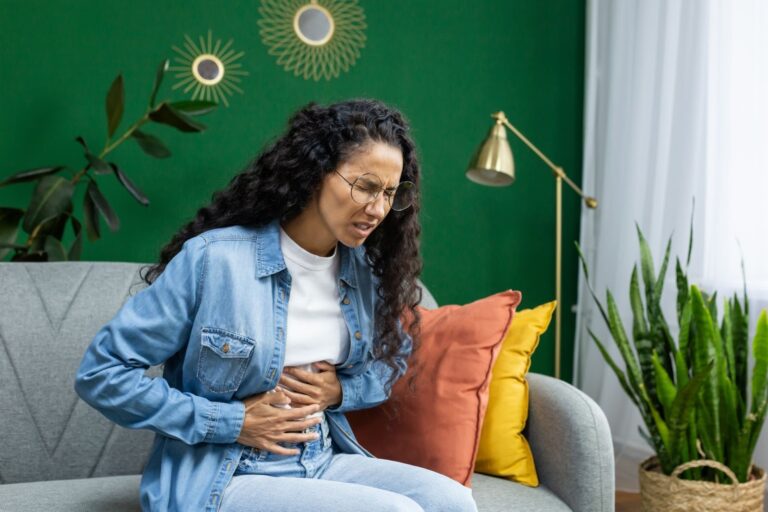 A woman clutches her stomach while sitting on a couch, showing signs of digestive discomfort
