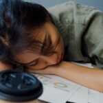 Exhausted woman asleep at a desk surrounded by paperwork and coffee cups, showing fatigue from a heavy workload