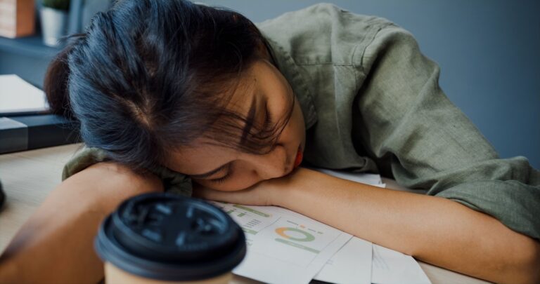 Exhausted woman asleep at a desk surrounded by paperwork and coffee cups, showing fatigue from a heavy workload