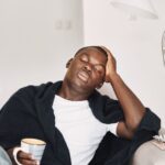 Tired African man seated on a couch, holding a cup of coffee while pressing his head, with pills beside him