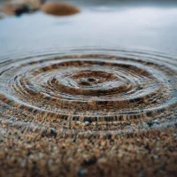 A high-angle, close-up shot of water ripples in a shallow puddle with a sandy bottom, creating an intricate pattern.