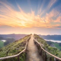 A winding dirt path with a rustic wooden fence leads up a grassy ridge with two calm lakes below, all under a vibrant sunset sky.